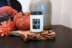 A white jar candle with a black lid and a black label reading 'BROWN BUTTER brulee' sits on a rustic wood slice, styled with orange pumpkins, autumn leaves, and a large ceramic vase in the background.