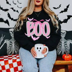 A woman wearing a black crewneck sweatshirt with a pink and white striped 'BOO' graphic and bat details, holding a white ghost-shaped mug while sitting on a bench with Halloween decorations.