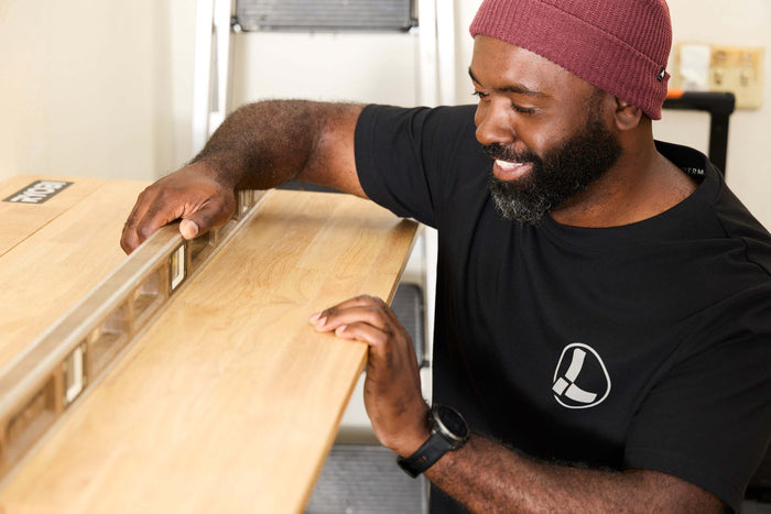 A person wearing a black Leatherman Classic Tee using a level tool to level a wooden shelf indoors.