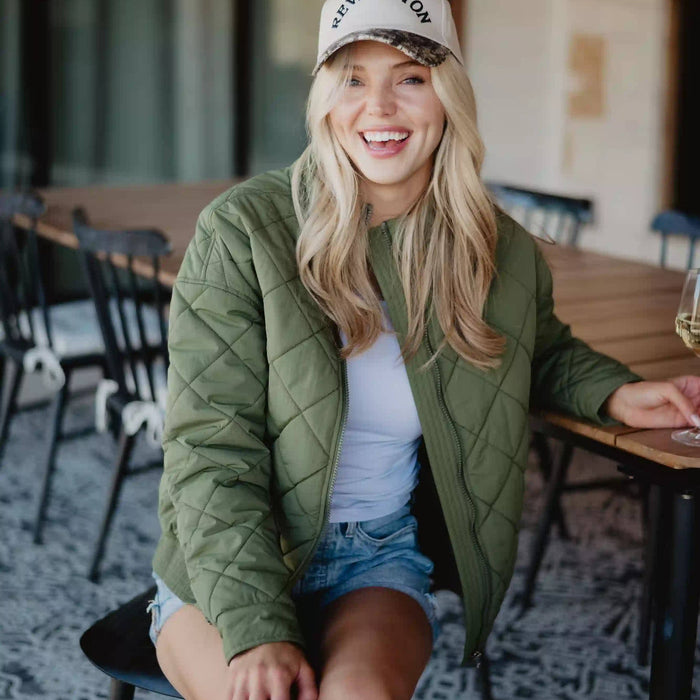 Woman sitting in a cafe wearing a green quilted jacket and white cap.
