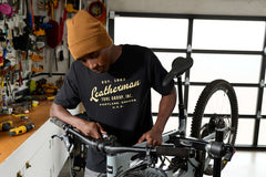 A person wearing a black Leatherman Vintage Tee working on a mountain bike in a garage workshop, surrounded by tools and equipment.