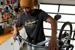 A person wearing a black Leatherman Vintage Tee working on a mountain bike in a garage workshop, surrounded by tools and equipment.
