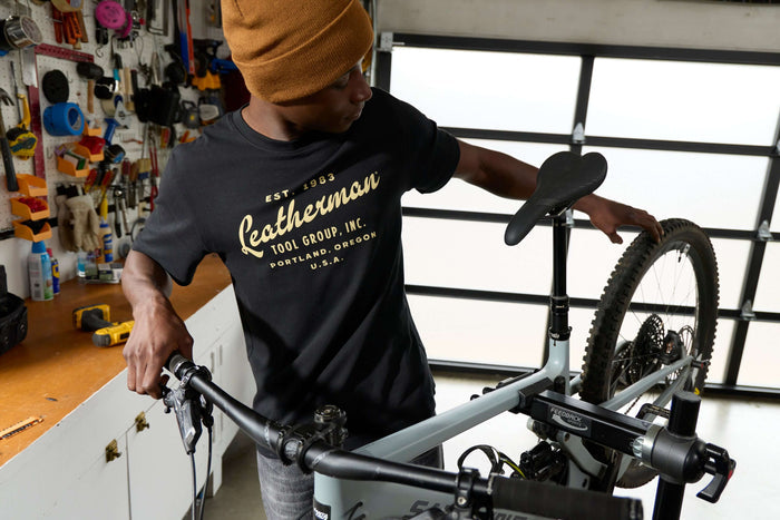 A person wearing a black Leatherman Vintage Tee working on a mountain bike in a garage workshop, surrounded by tools and equipment.